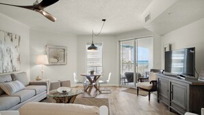 A spacious living room with a large TV, a dining area with a glass table, and a balcony with a view of the ocean. - Dunes Of Seagrove (Santa Rosa Beach)