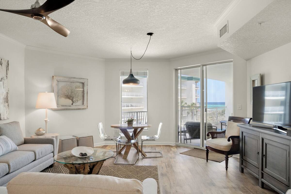 A spacious living room with a large TV, a dining area with a glass table, and a balcony with a view of the ocean.