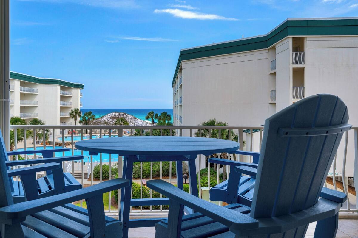 A balcony with blue Adirondack chairs and a round table, overlooking a pool and a building with a green roof.