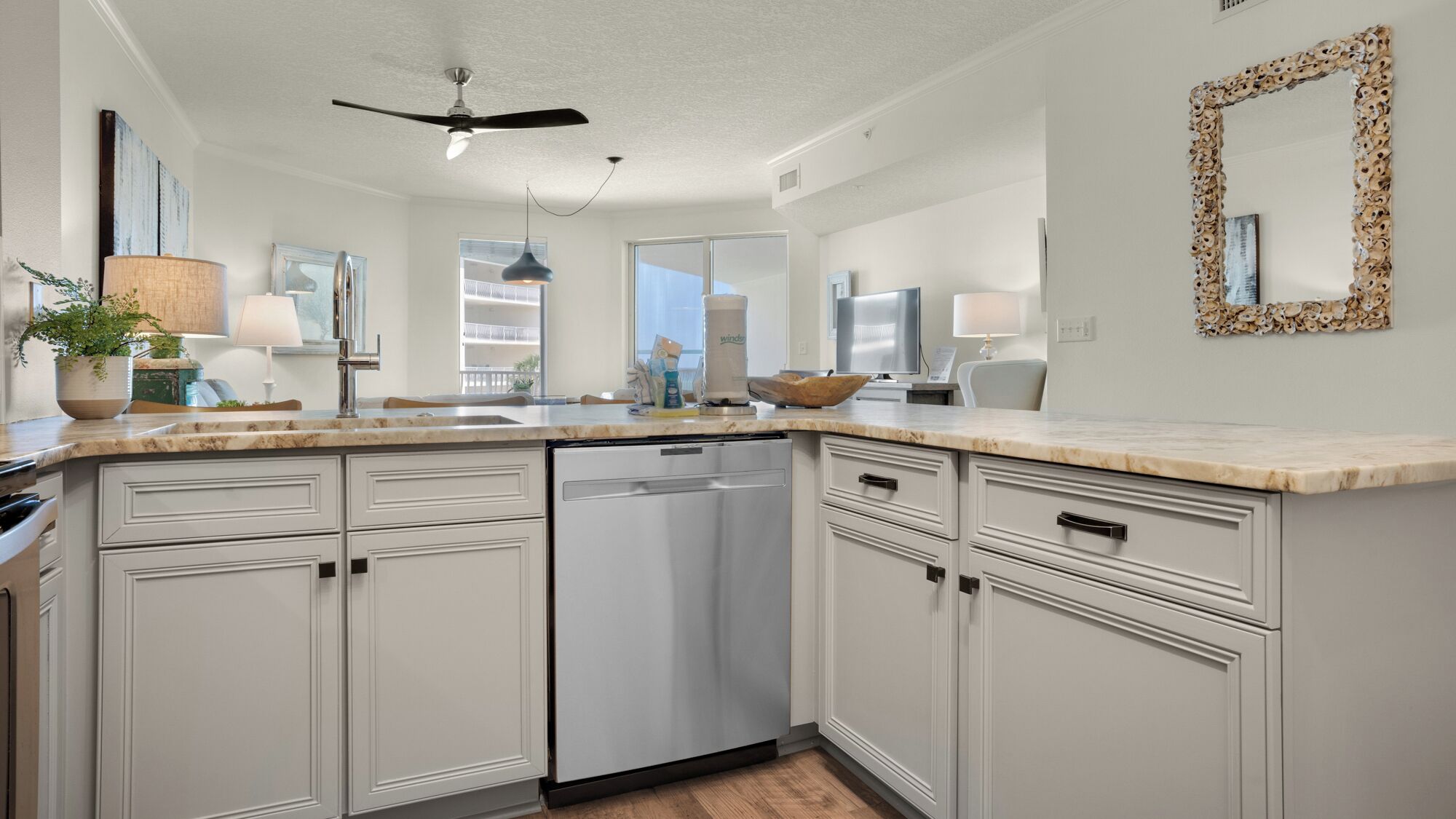 A modern kitchen with white cabinetry, a built-in dishwasher, a ceiling fan, and a large window providing natural light.