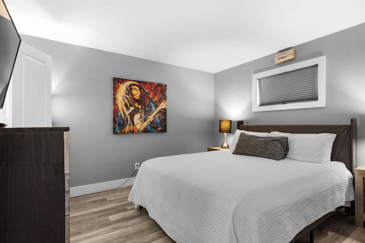 A bedroom with a bed, two bedside lamps, a painting, a window with blinds, and a wooden nightstand.