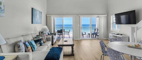 A spacious living room with a sofa, coffee table, and dining area. A large TV is mounted on the wall, and there is a balcony with a view of the ocean.