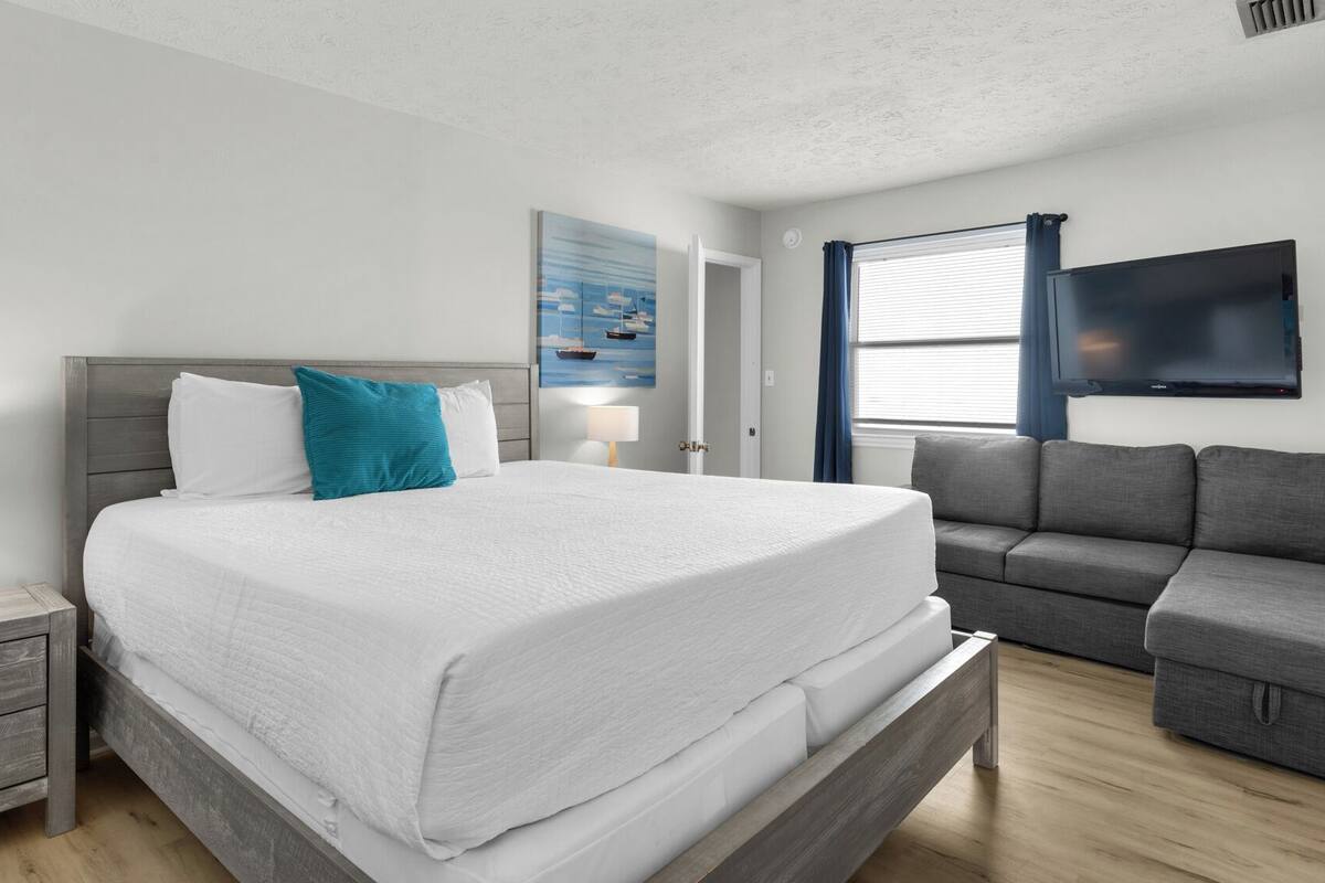 A neatly arranged bedroom with a bed, bedside tables, a grey sofa, and a TV.