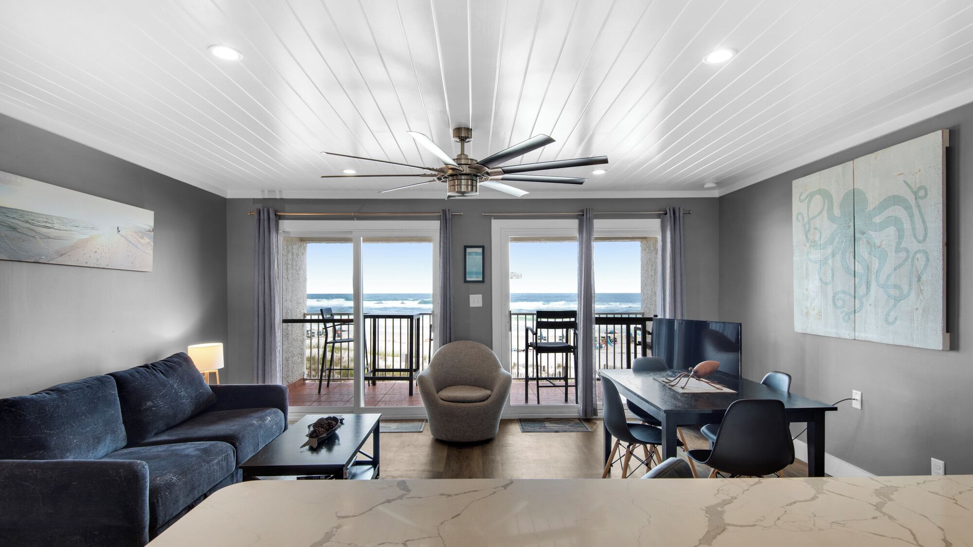 A modern living room with a sofa, coffee table, and a dining area with chairs and a table. There is a ceiling fan, a painting on the wall, and a view of the ocean through large windows.