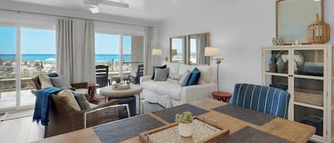 Een ruime woonkamer met een groot raam met uitzicht op een strand, een eethoek met een houten tafel, en een zithoek met een bank en fauteuils.
