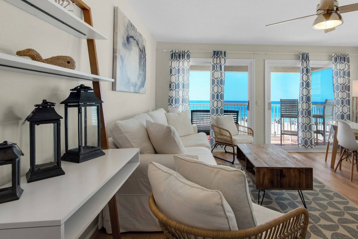 A modern living room with a white sofa, wicker chairs, a dining table, and a view of the ocean through large windows.