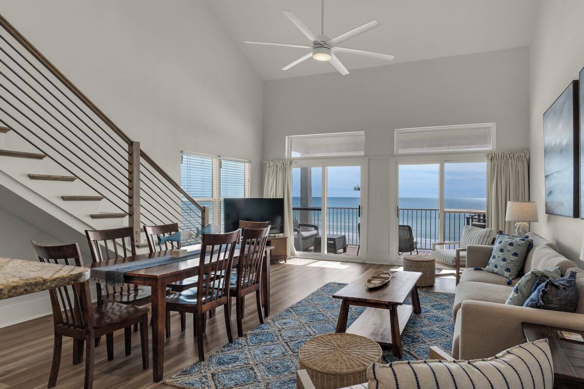 A spacious living room with a dining area, a ceiling fan, a large window with a view of the sea, and a balcony with outdoor furniture.