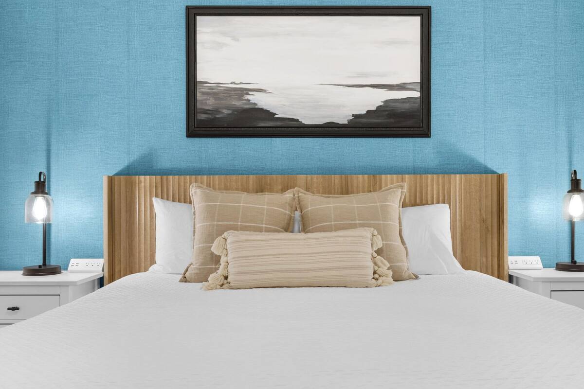 A neatly made bed with a white comforter and beige pillows, flanked by two bedside tables with lamps, against a blue wall with a framed picture above.
