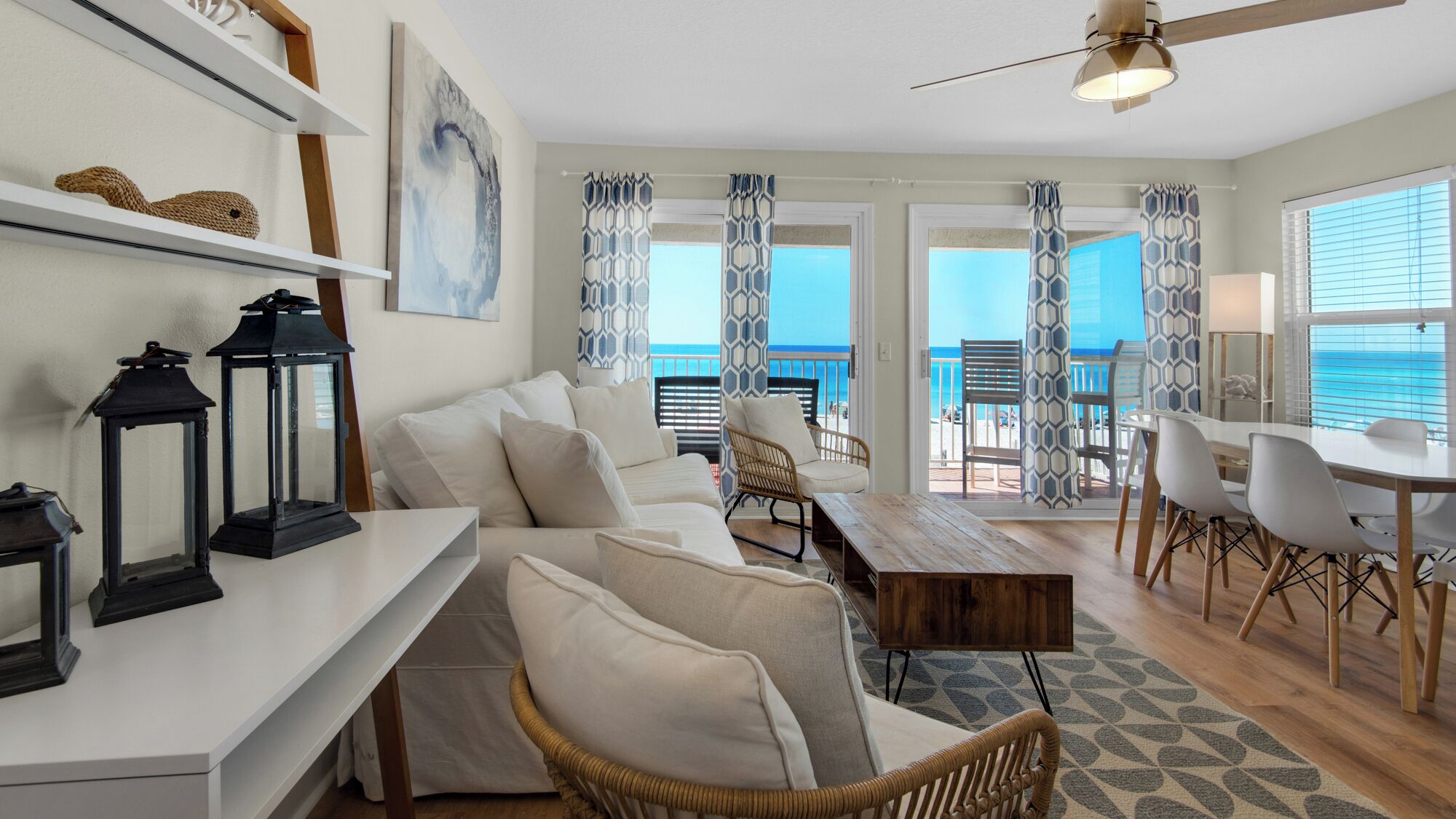 A modern living room with a white sofa, wicker chairs, a dining table, and a view of the ocean through large windows.
