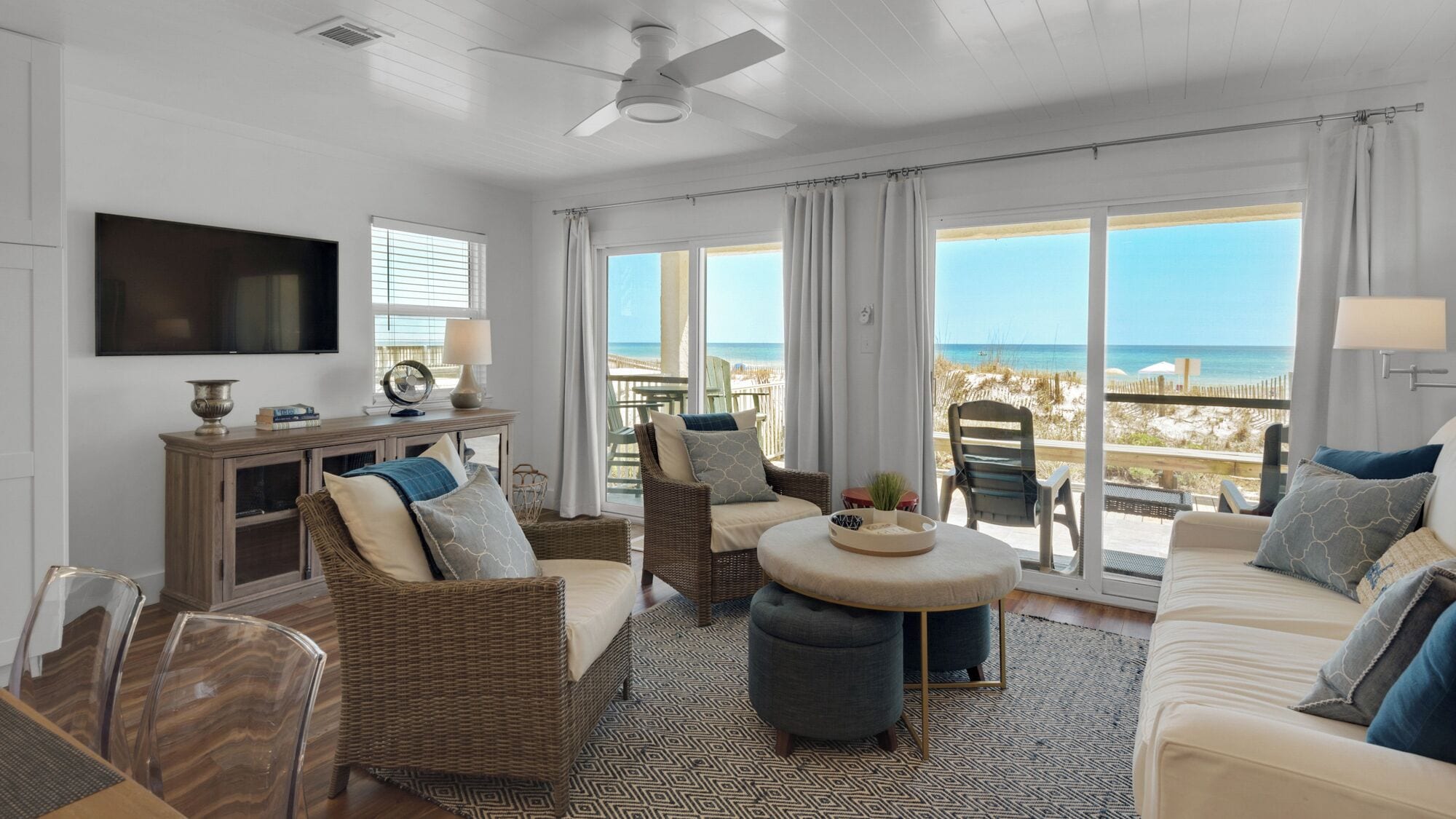 A spacious living room with a view of the beach, furnished with wicker chairs, a sofa, and a round table.