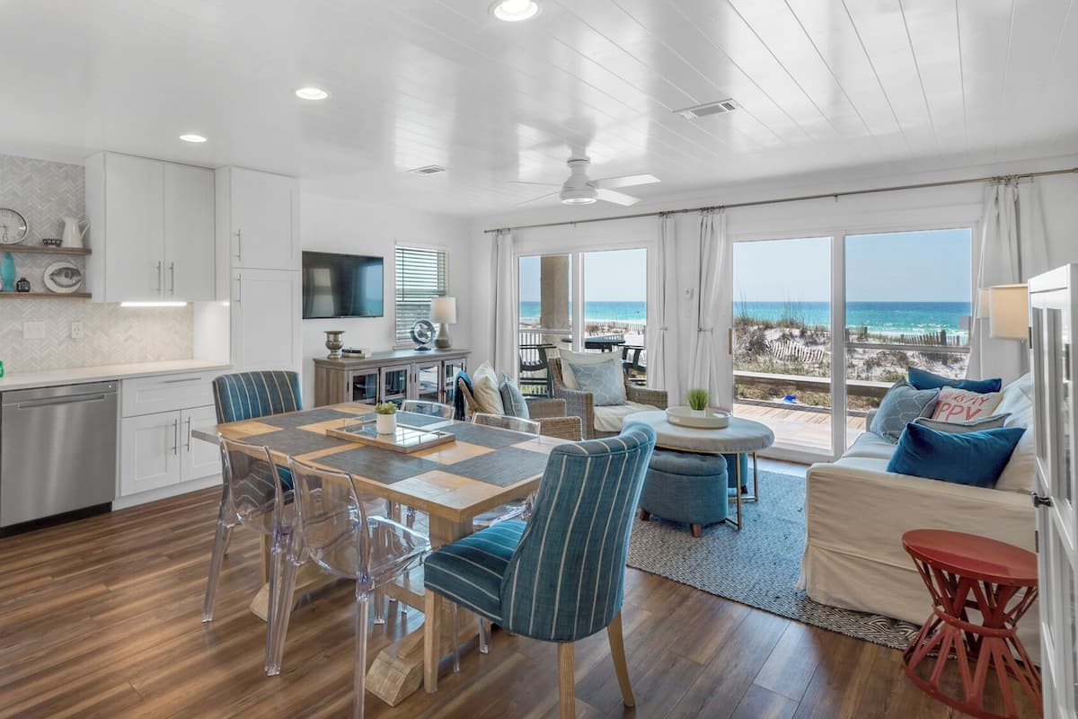 A spacious kitchen with modern appliances, a dining area with clear chairs, and a living area with a view of the beach.