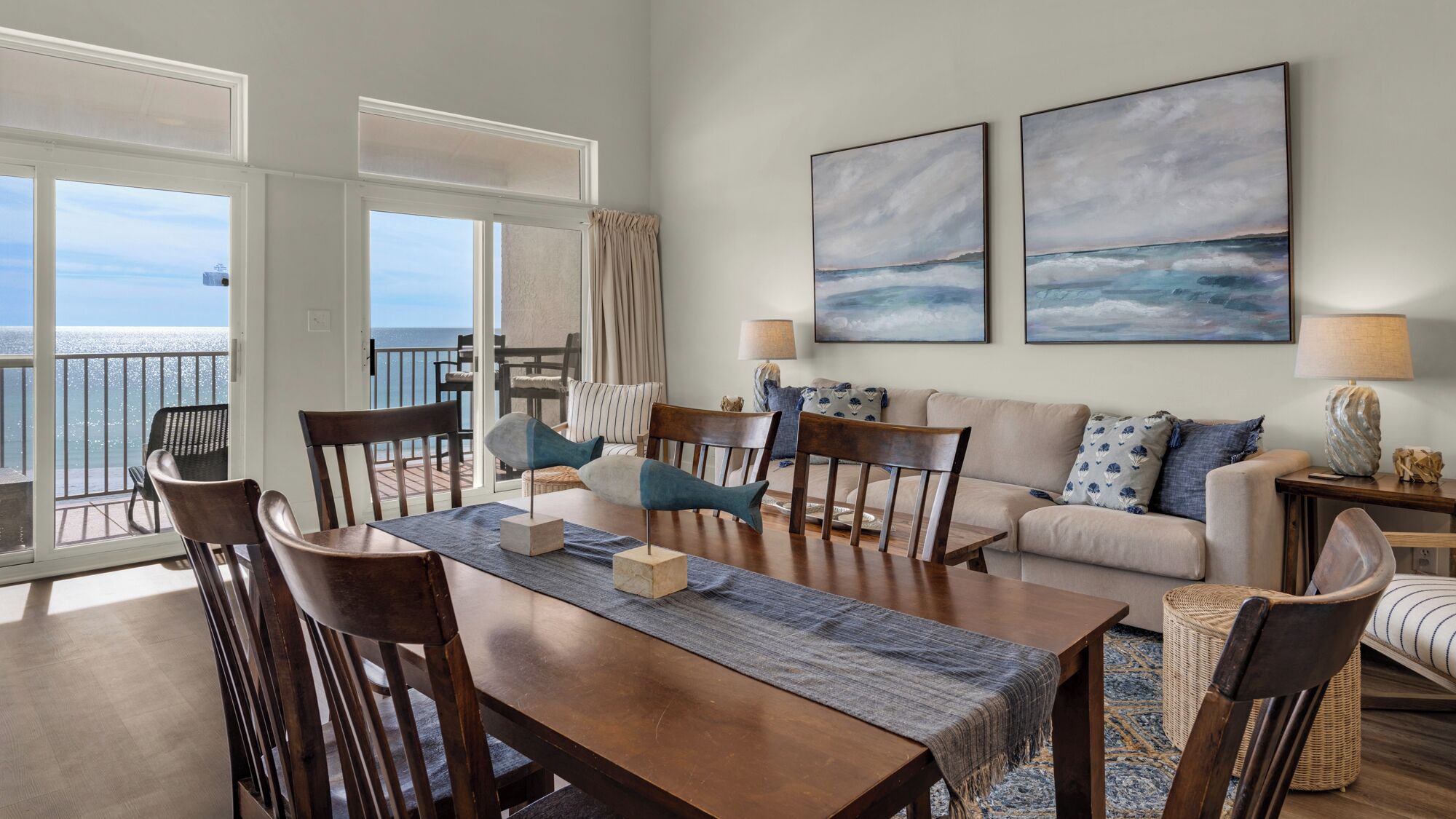 Een ruime woonkamer met een eettafel, stoelen en een bank. Twee grote schilderijen hangen aan de muur, een balkon met uitzicht op zee, en een lamp op een bijzettafeltje.