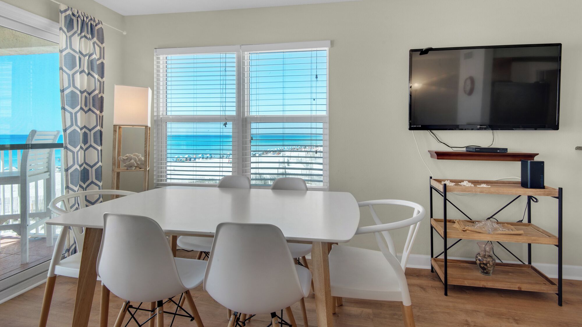 A dining area with a white table and chairs, a TV on a wall, and a view of the beach through large windows.
