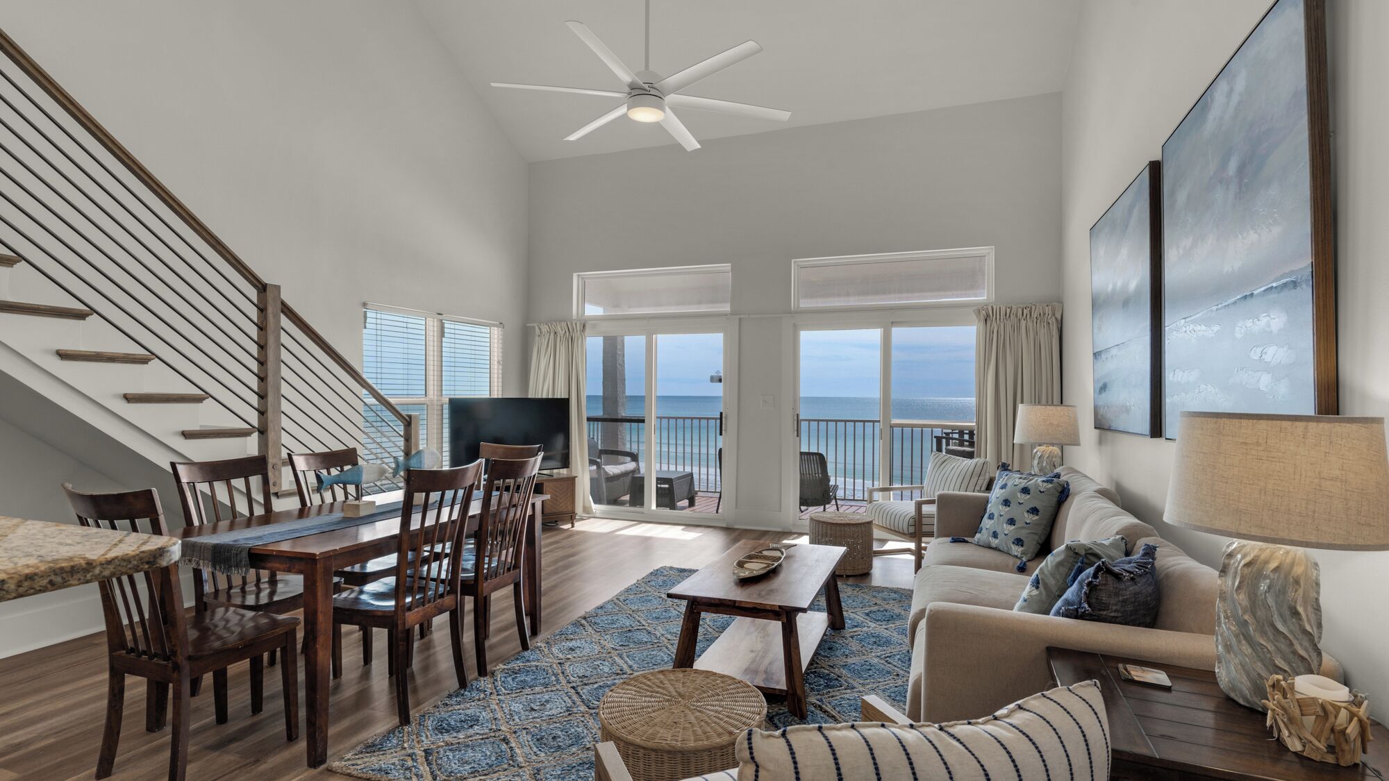 A spacious living room with a dining area, a ceiling fan, a large window with a view of the sea, and a balcony with outdoor furniture.