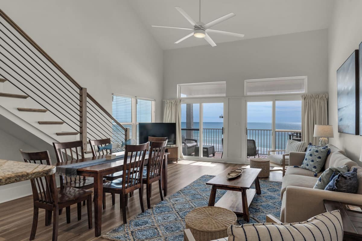 A spacious living room with a dining area, a ceiling fan, a large window with a view of the sea, and a balcony with outdoor furniture.