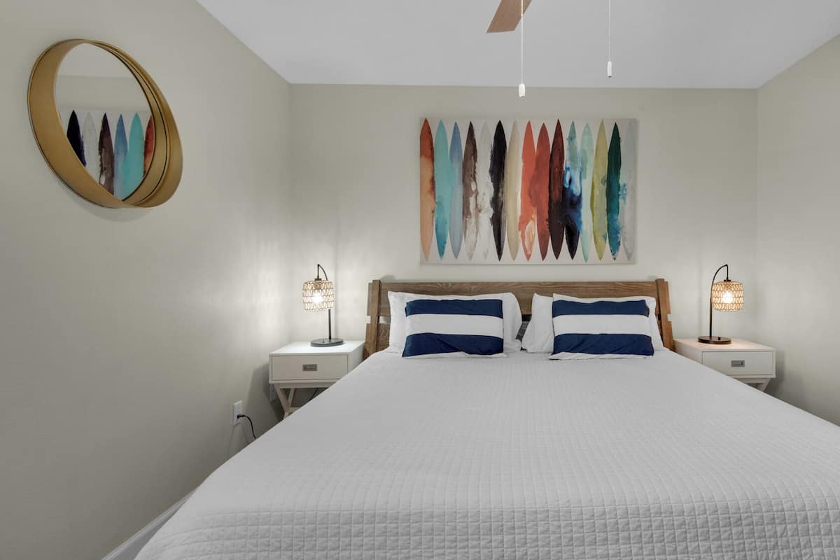 A bedroom with a large bed, two bedside tables, a ceiling fan, a wall-mounted mirror, and a colorful abstract painting.