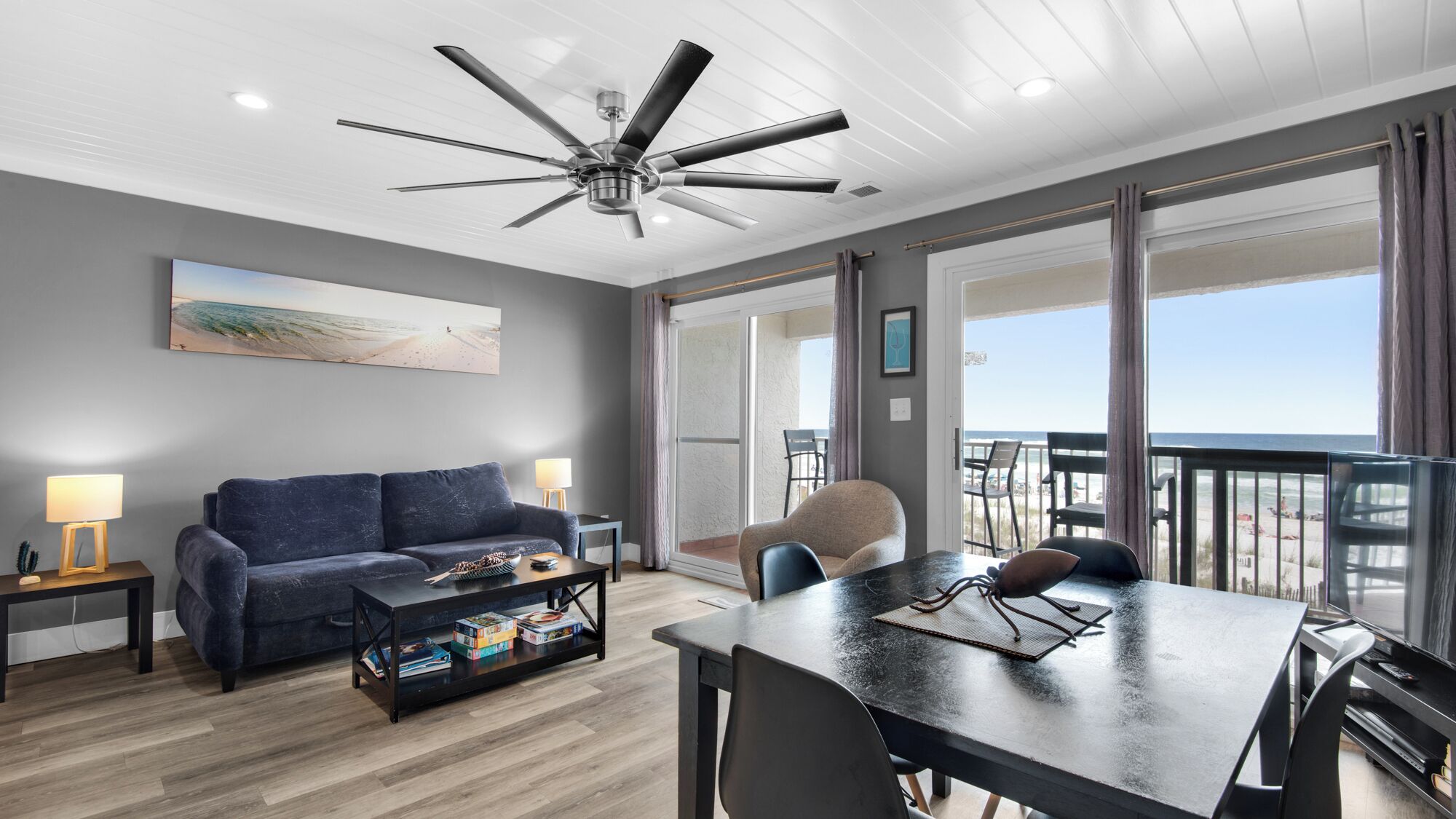 A modern living room with a ceiling fan, a sofa, a coffee table with books, a dining table with chairs, and a large window with a view of the beach.
