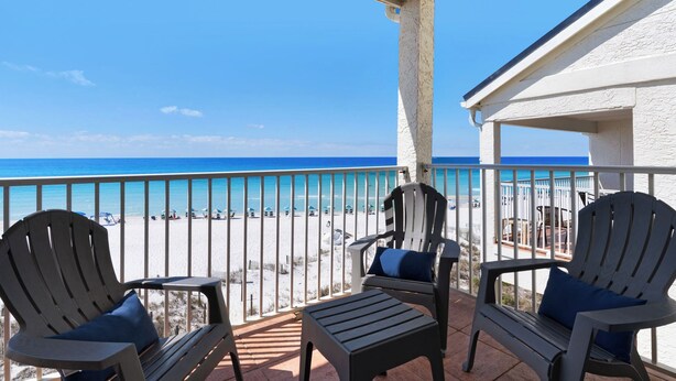 Un balcon avec deux fauteuils Adirondack et une petite table, donnant sur une plage parsemée de parasols et animée par la présence de nombreuses personnes.