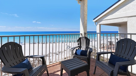 A balcony with two Adirondack chairs and a small table, overlooking a beach with umbrellas and people.
