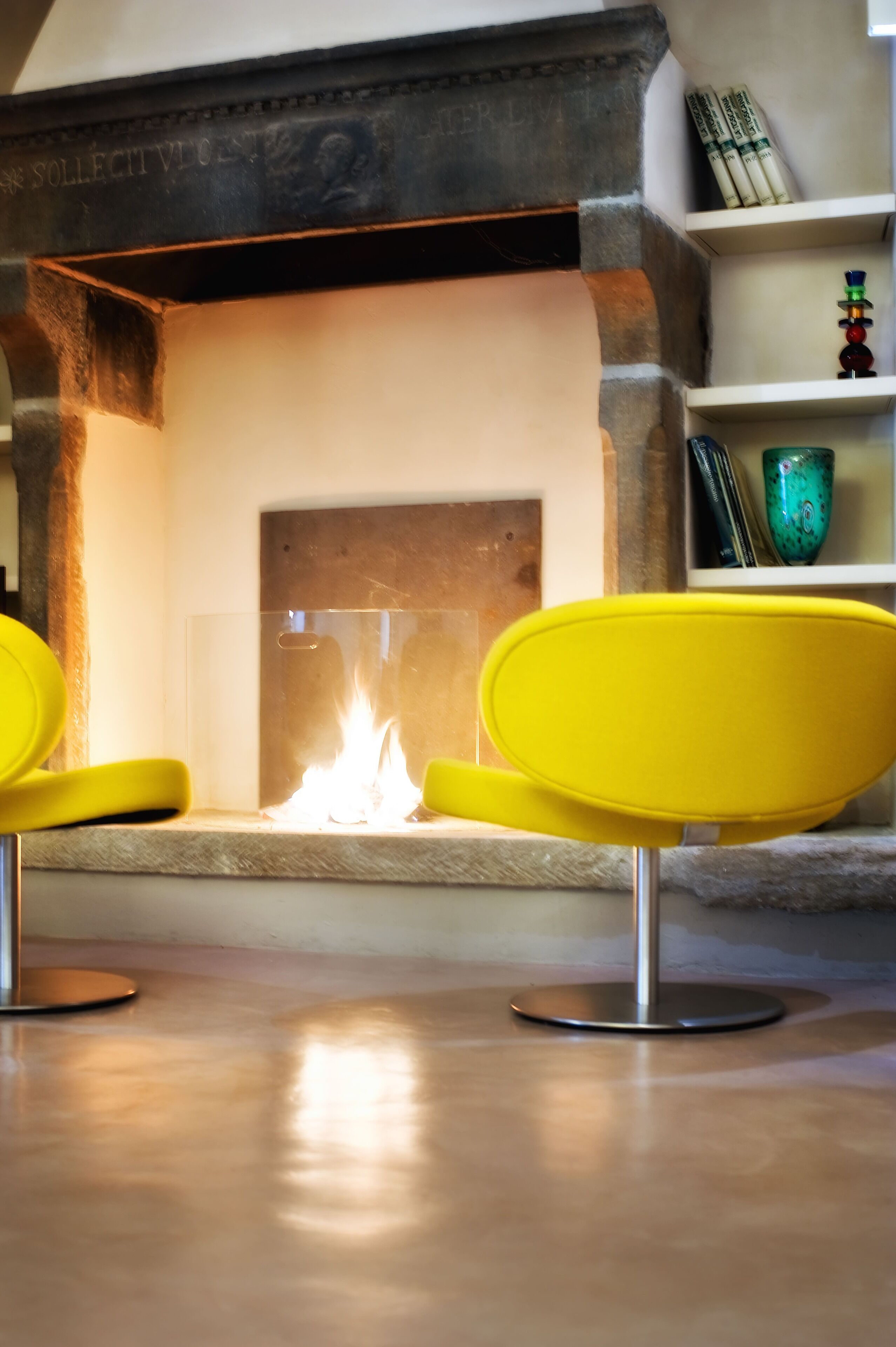 Une cheminée avec une façade en verre transparent, deux fauteuils jaunes placés devant, et une cheminée en pierre au-dessus.