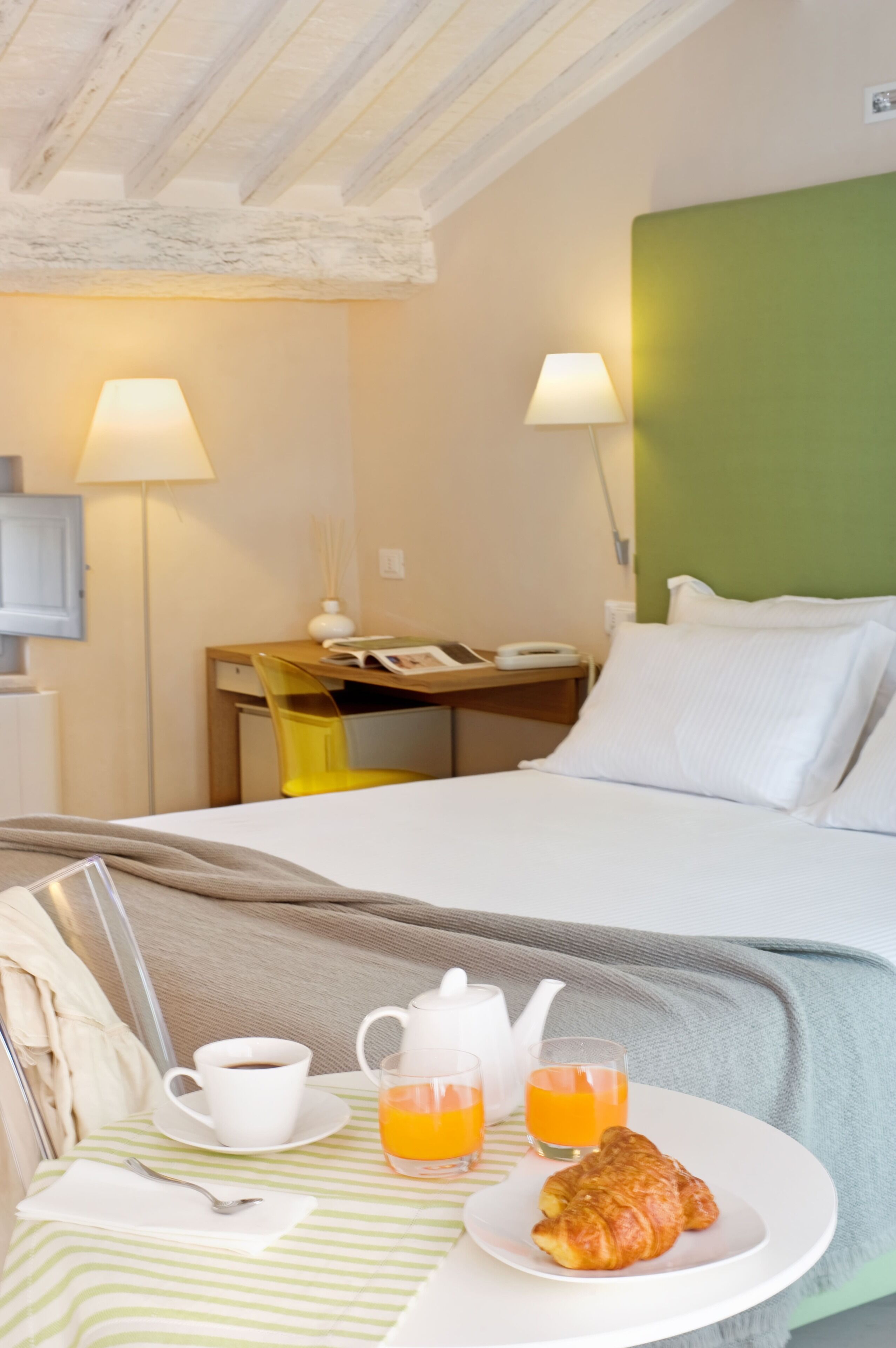 A hotel room with a bed, bedside table, and a breakfast setup including a cup of coffee, a teapot, glasses of orange juice, and a croissant.