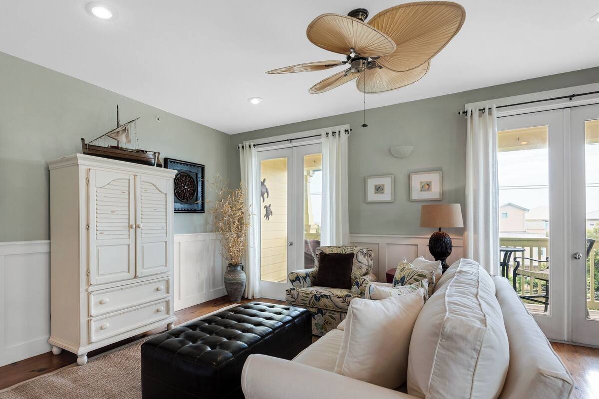 A living room with a white sofa, a black ottoman, a ceiling fan, and a view of the outside.