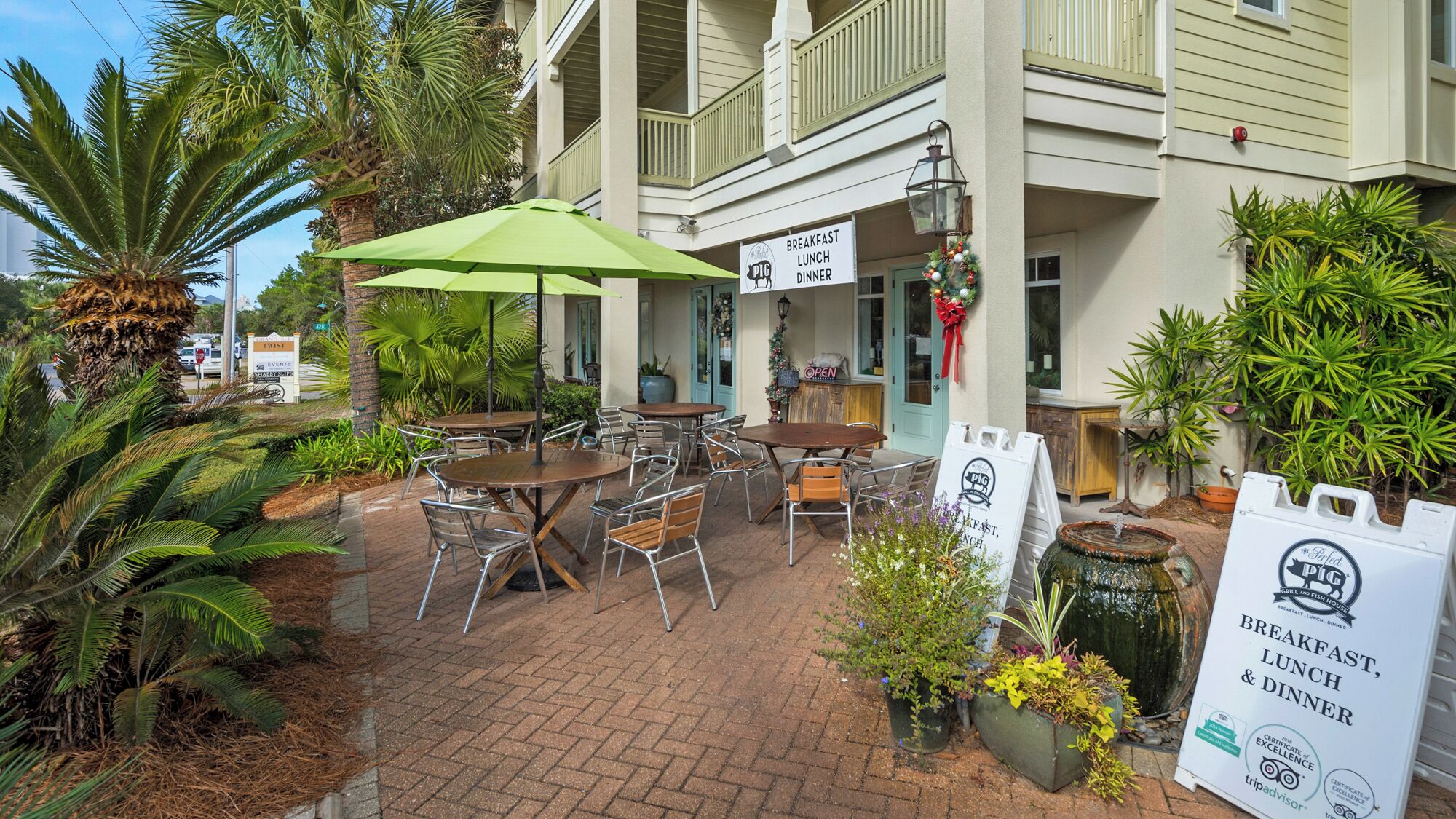 An outdoor patio with tables and chairs, a green umbrella, and a sign for a restaurant offering breakfast, lunch, and dinner.