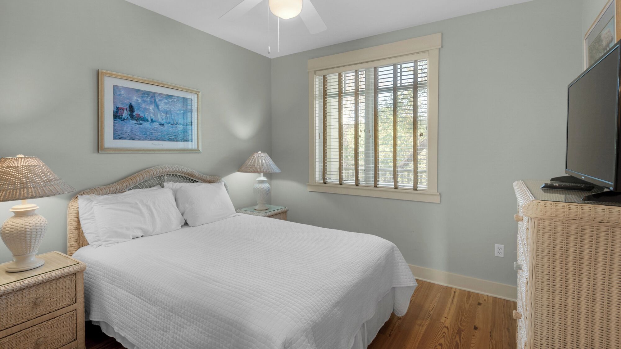 Une chambre à coucher avec un lit, deux lampes de chevet, un tableau, une fenêtre avec des stores et un téléviseur posé sur un meuble.