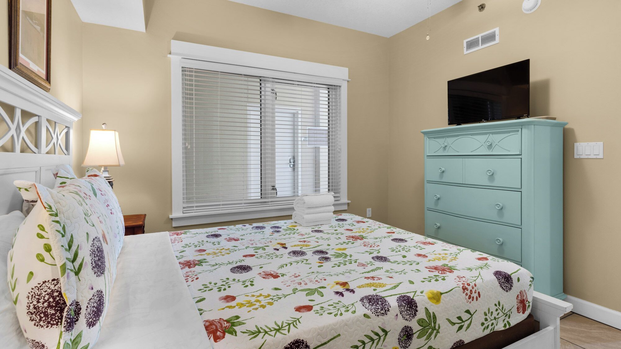 Un dormitorio con cama, cómoda, ventana con persianas, lámpara y un cuadro en la pared.
