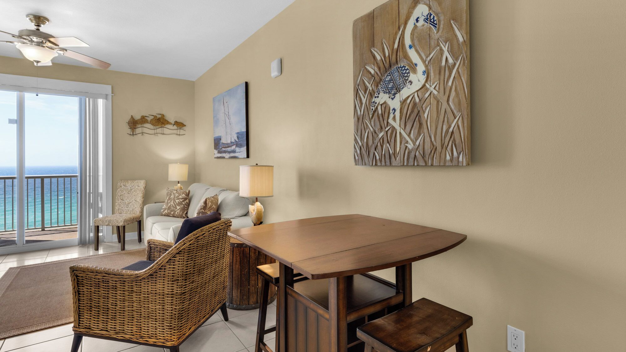 A living room with a view of the ocean, a dining table, wicker chairs, a sofa, and artwork on the walls.