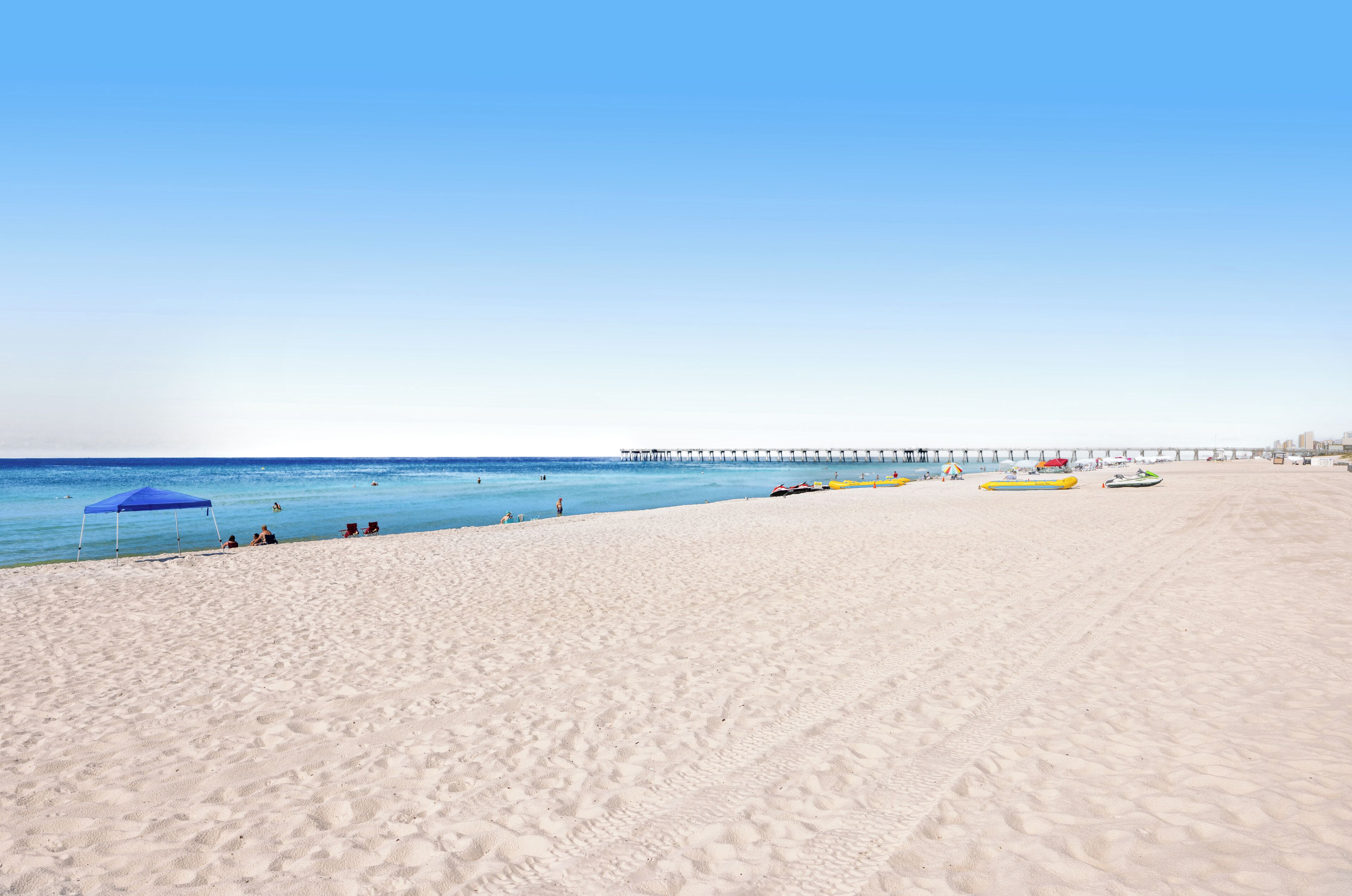 Une plage avec une jetée qui s’avance dans l’eau, un ciel dégagé, quelques parasols et quelques bateaux.