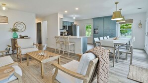 Un vaste espace de vie comprenant une table basse en bois, des canapés blancs et une table à manger avec des chaises. On aperçoit au fond une cuisine avec des meubles bleus et un réfrigérateur.