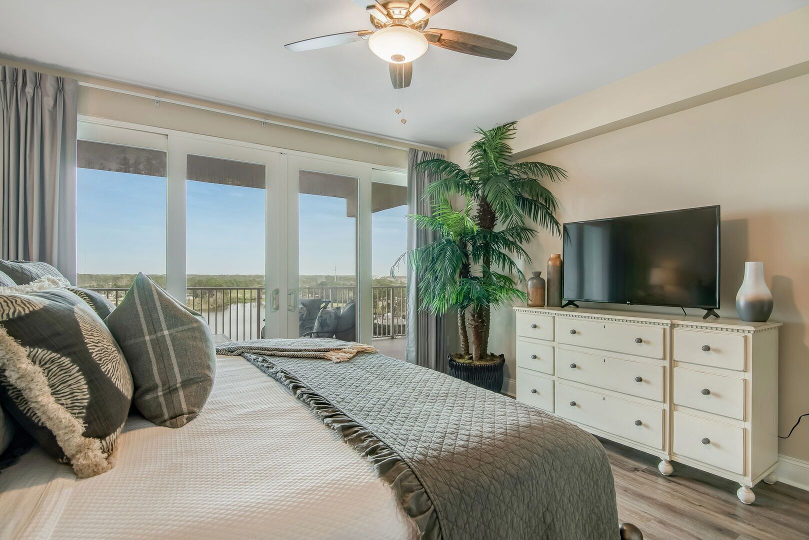 A bedroom with a large bed, a ceiling fan, a TV, a dresser, and a view of the outdoors.