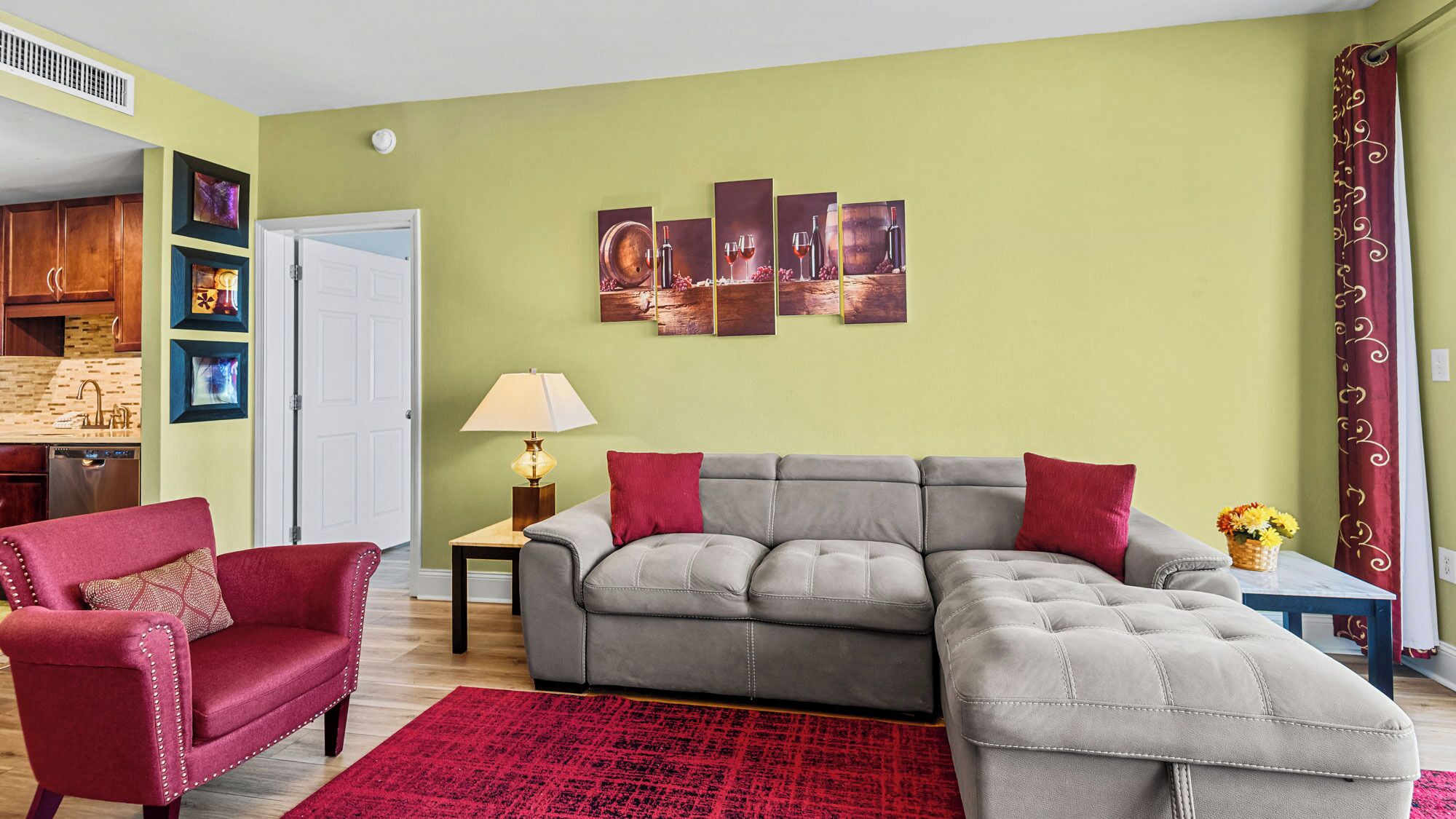 A living room with a grey sectional sofa, red armchairs, a red rug, and a wall with framed artwork.