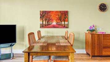 Un comedor con una mesa de madera, seis sillas, un cuadro de un sendero bordeado de árboles, un reloj, un televisor y un mueble de madera con cajones.