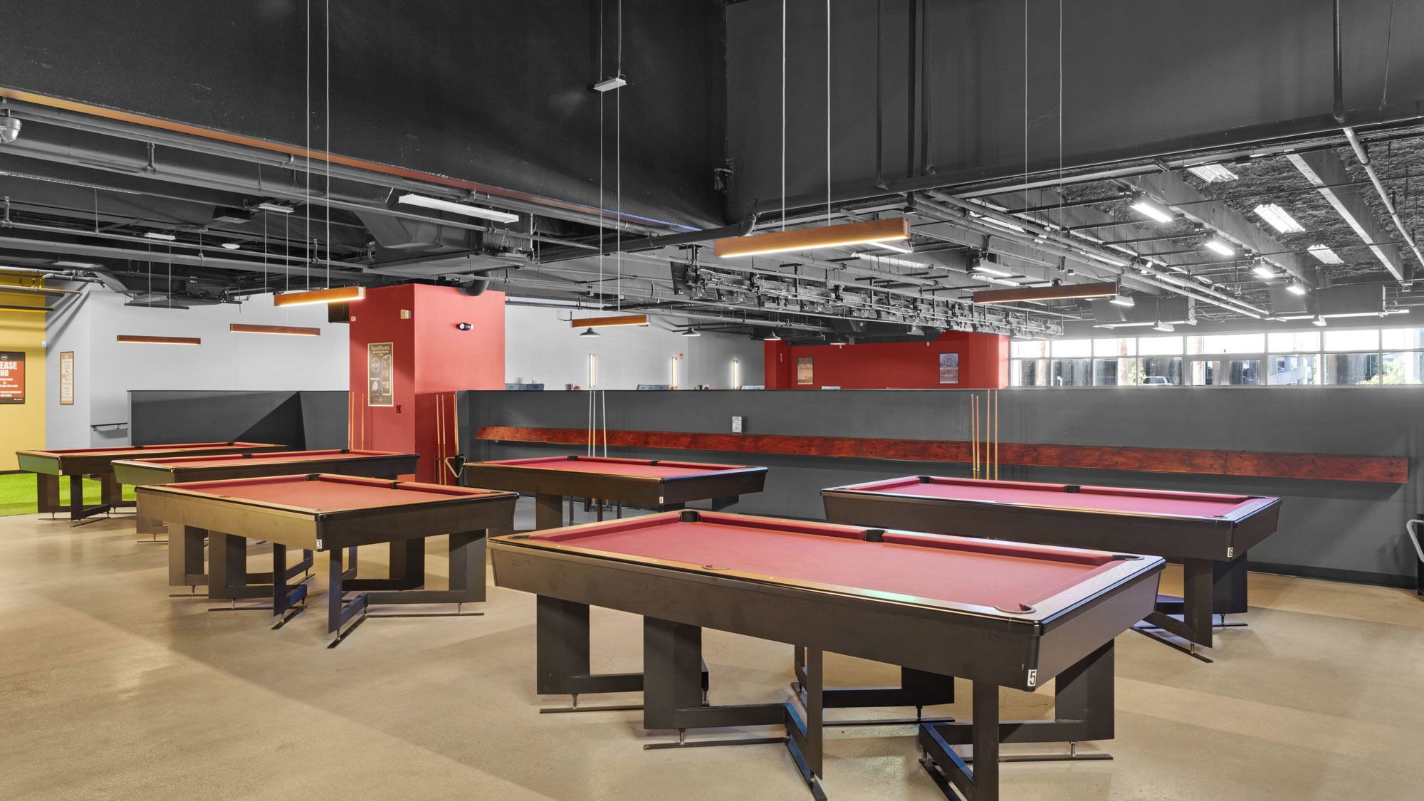 A billiards hall with multiple red pool tables, a red machine, and a grey wall with windows.