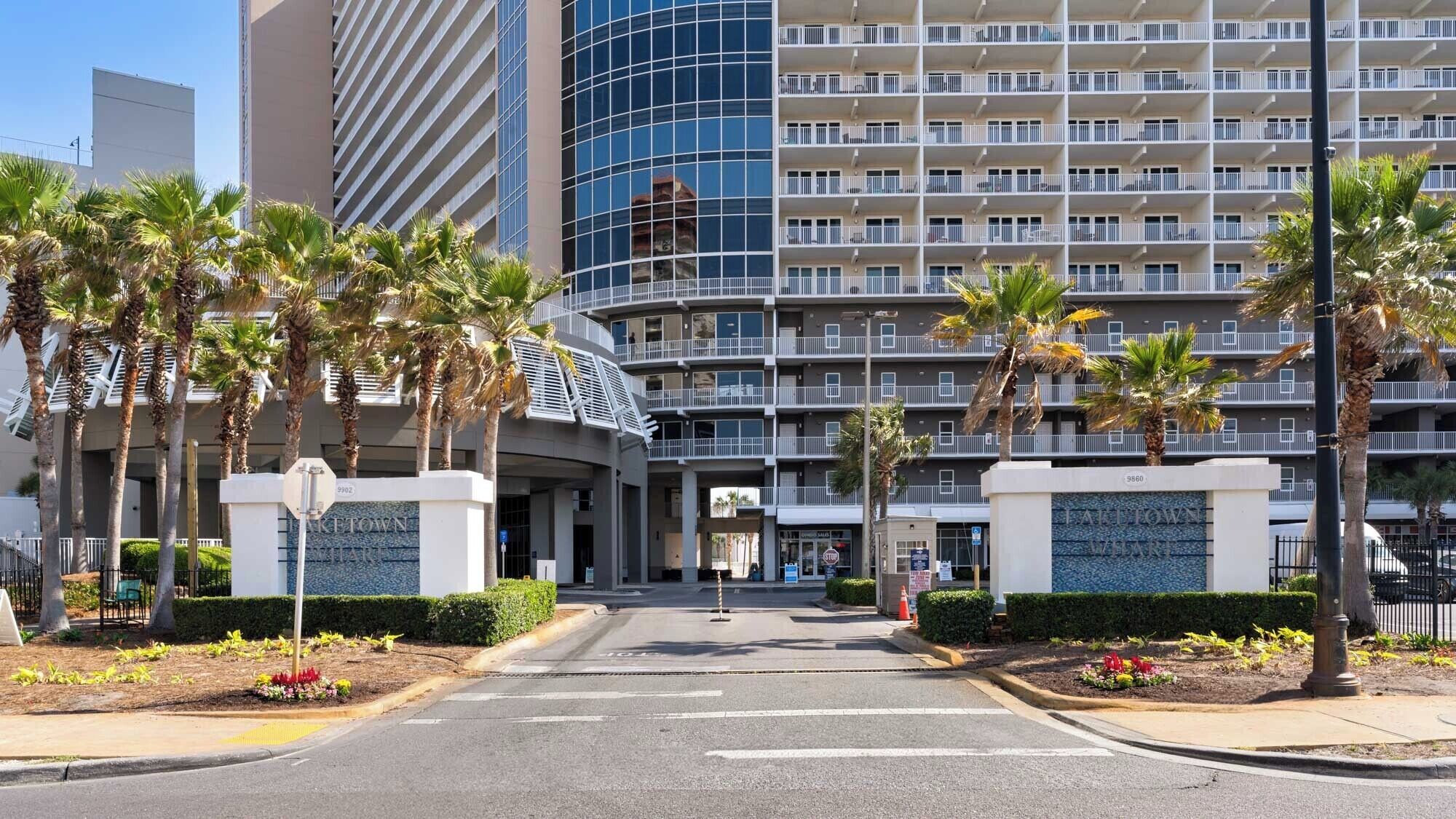 Une entrée d’hôtel moderne avec une façade incurvée en verre, des palmiers et un ciel dégagé.