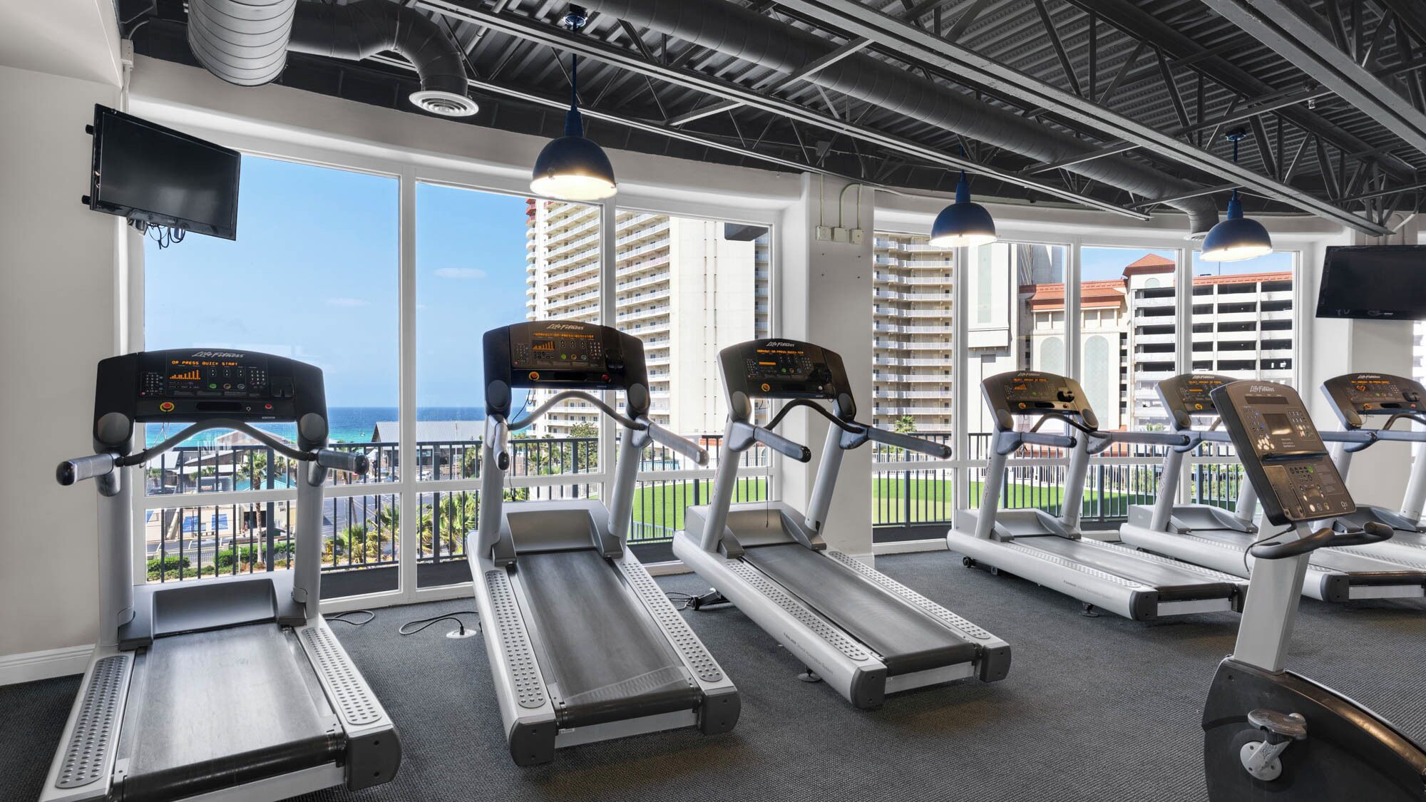 A gym with treadmills and exercise equipment, large windows with a view of a building and a body of water.
