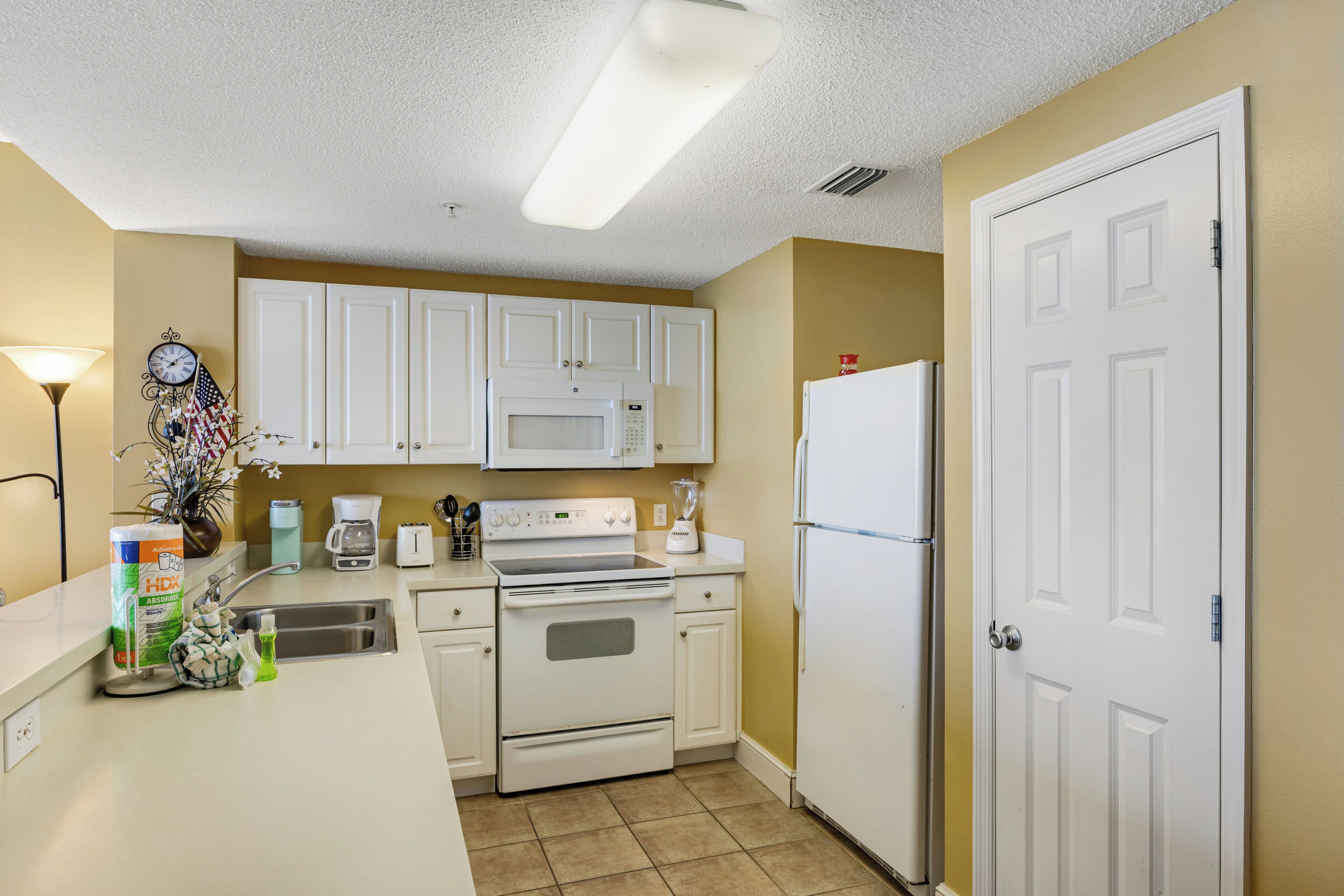 A small kitchen with white cabinets, a microwave, stove, and refrigerator. There is a clock on the wall and a plant on the counter.