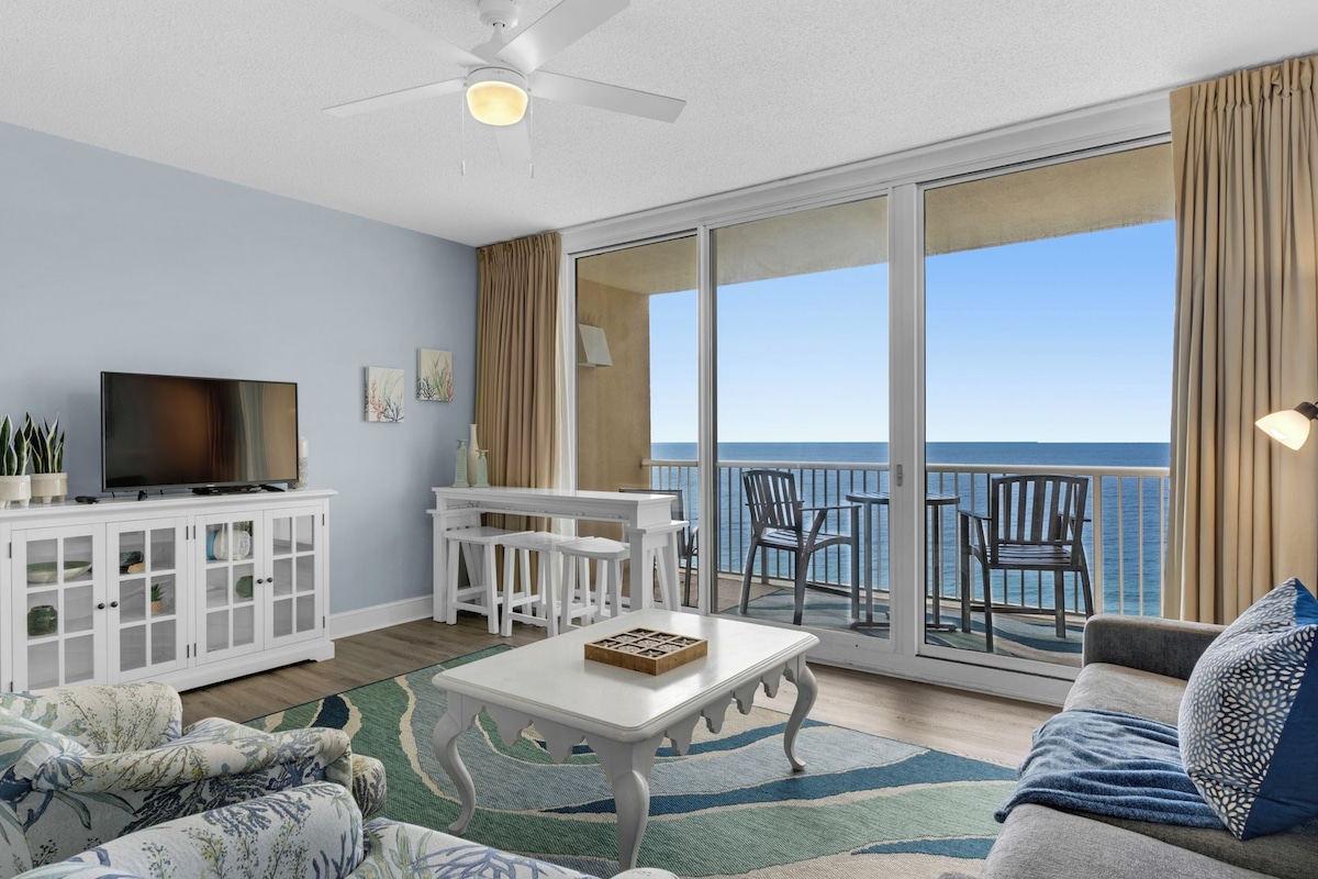 A living room with a TV, white furniture, a coffee table, and a view of the ocean.