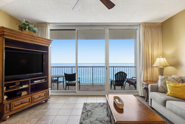 A living room with a large flat-screen TV, a wooden entertainment center, a sofa, a coffee table, and a balcony with a view of the ocean.