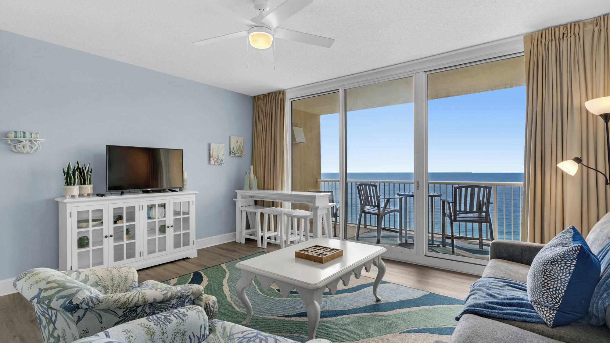 A living room with a TV, white furniture, a coffee table, and a view of the ocean.