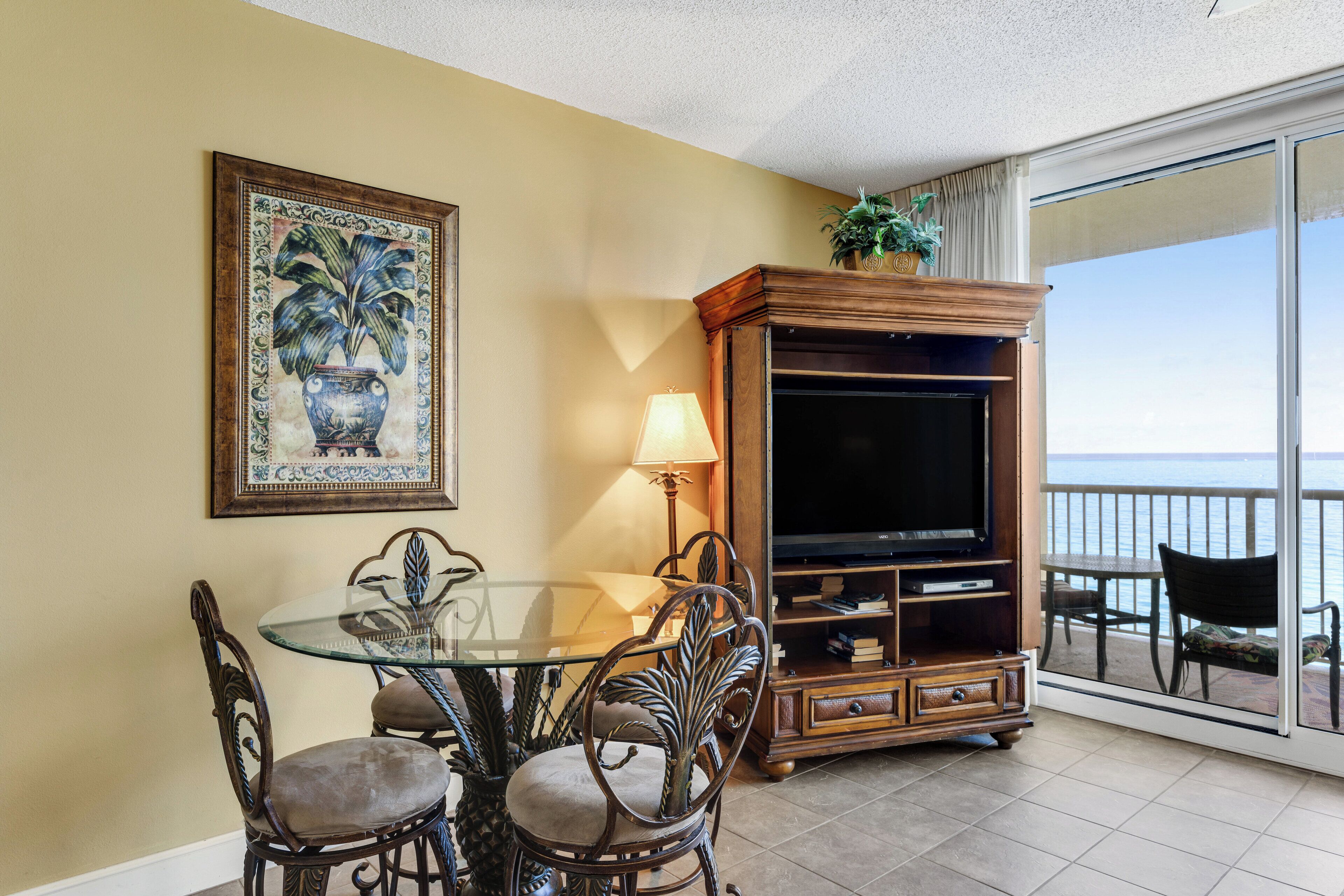 A dining area with a glass table and chairs, a television on a wooden stand, and a balcony with a view of the sea.