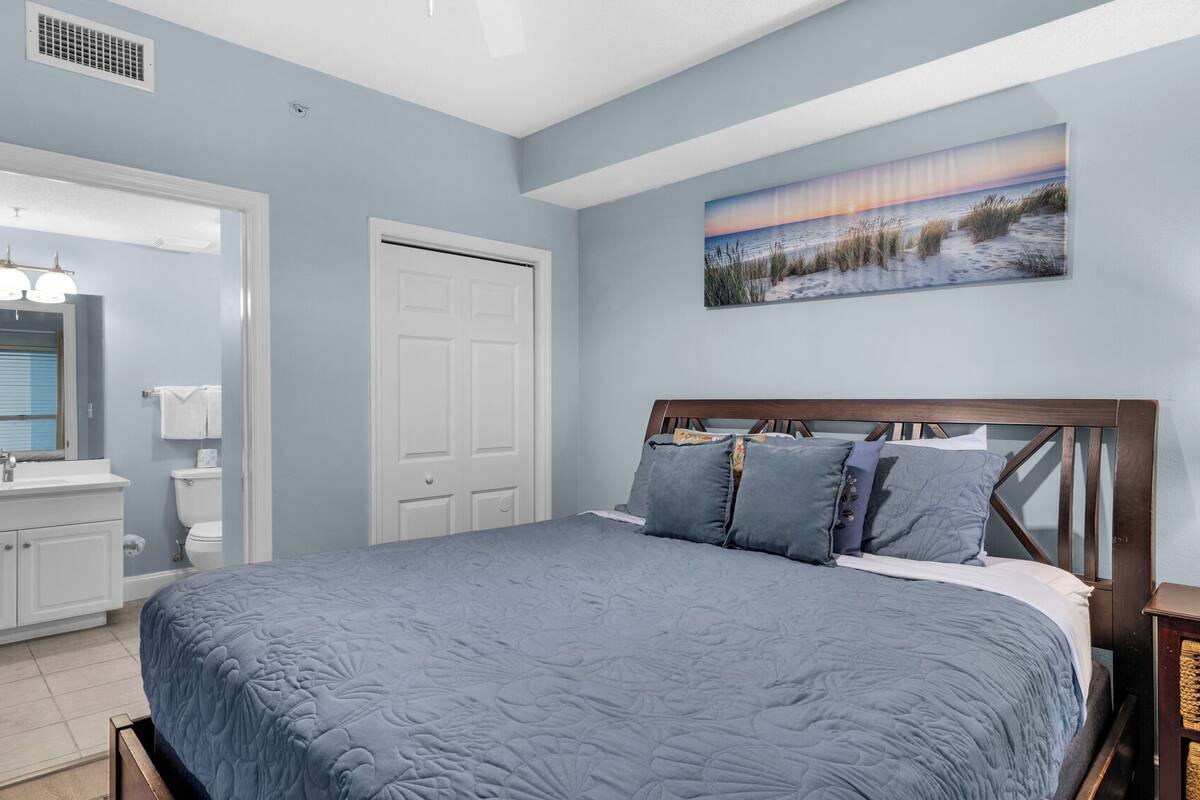 A bedroom with a large bed, a nightstand, a lamp, a painting of a beach scene, and a bathroom with a mirror and sink.
