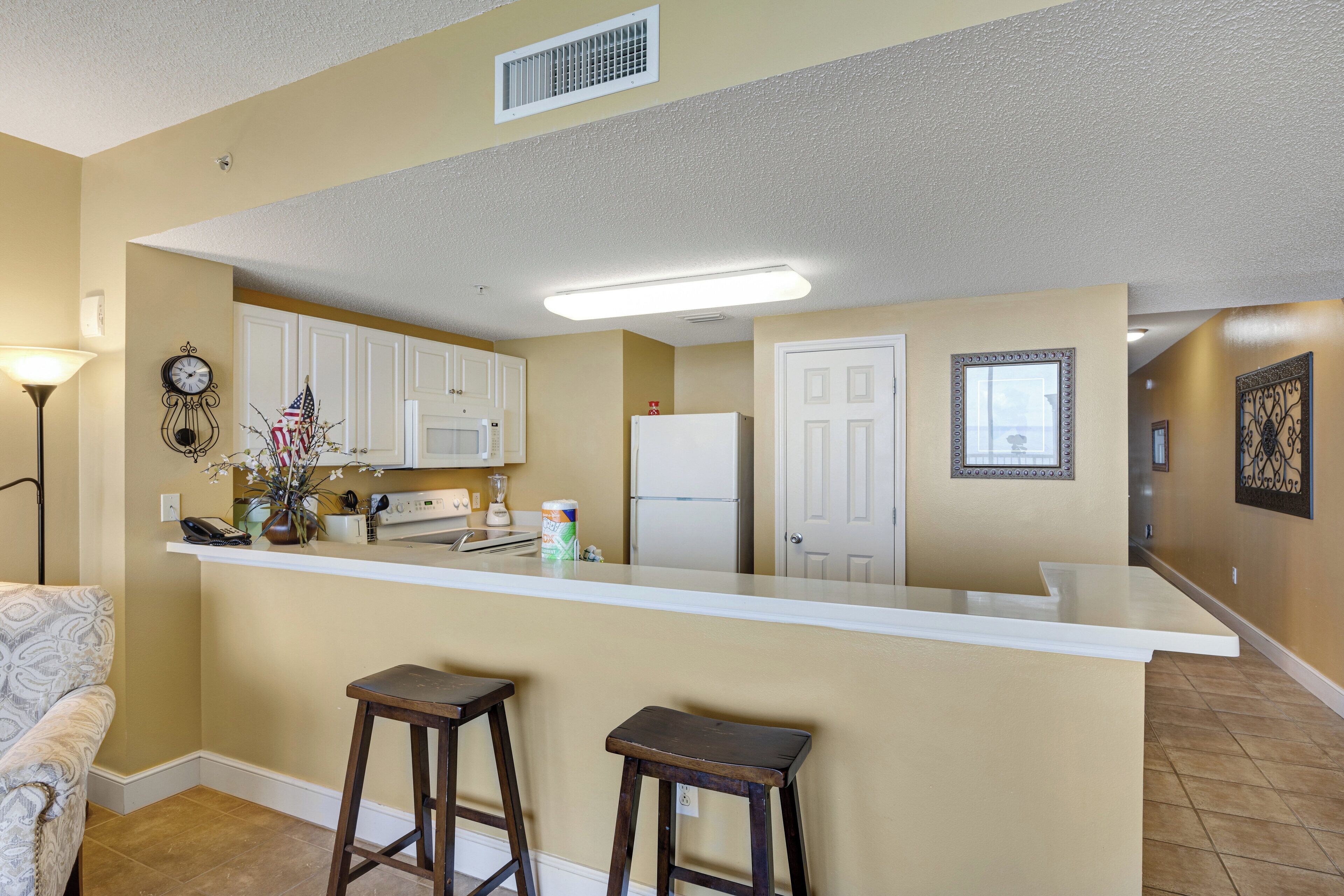A kitchen with a central island, white cabinets, and a microwave. There are two stools at the island, and a clock on the wall.