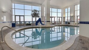 Una piscina interior con paredes de vidrio transparente, una silla azul y una palmera afuera.