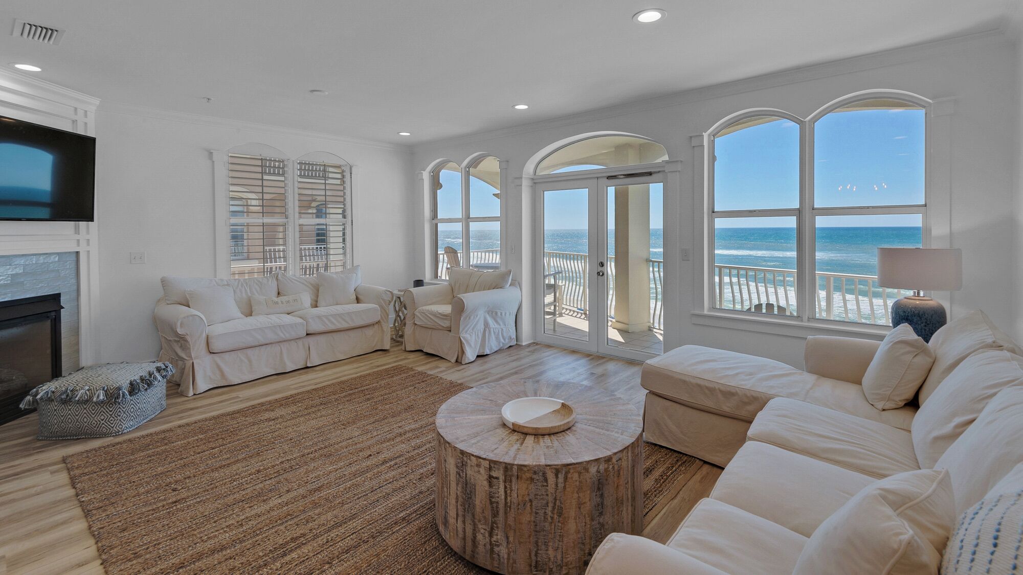 A spacious living room with a fireplace, white sofas, a round coffee table, and large windows overlooking the ocean.