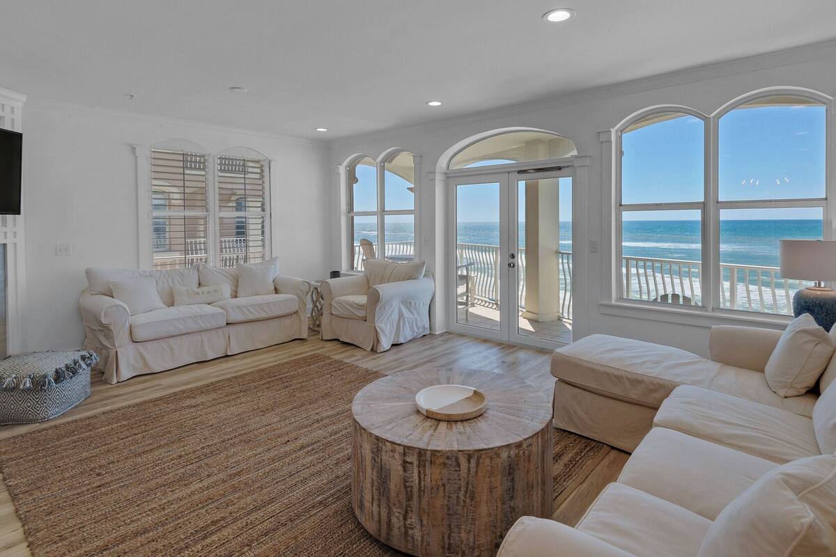 A spacious living room with a fireplace, white sofas, a round coffee table, and large windows overlooking the ocean.