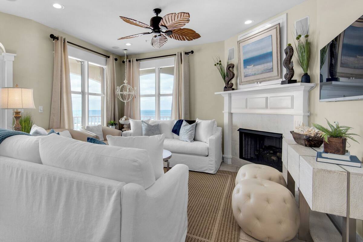 A spacious living room with a white fireplace, large windows, and a ceiling fan.