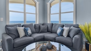 A grey sectional sofa with patterned cushions, a glass coffee table, and a view of the ocean through large windows. - Monterey Condos (Panama City Beach)